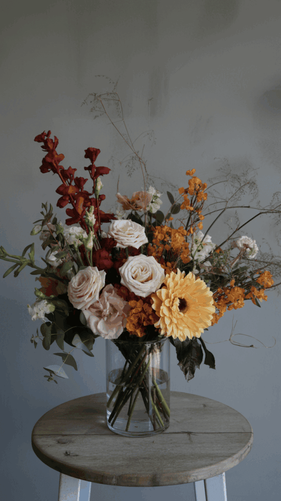 A bouquet of red, pink, and orange flowers with greenery arranged in a transparent glass vase on a wooden stool against a plain background.
