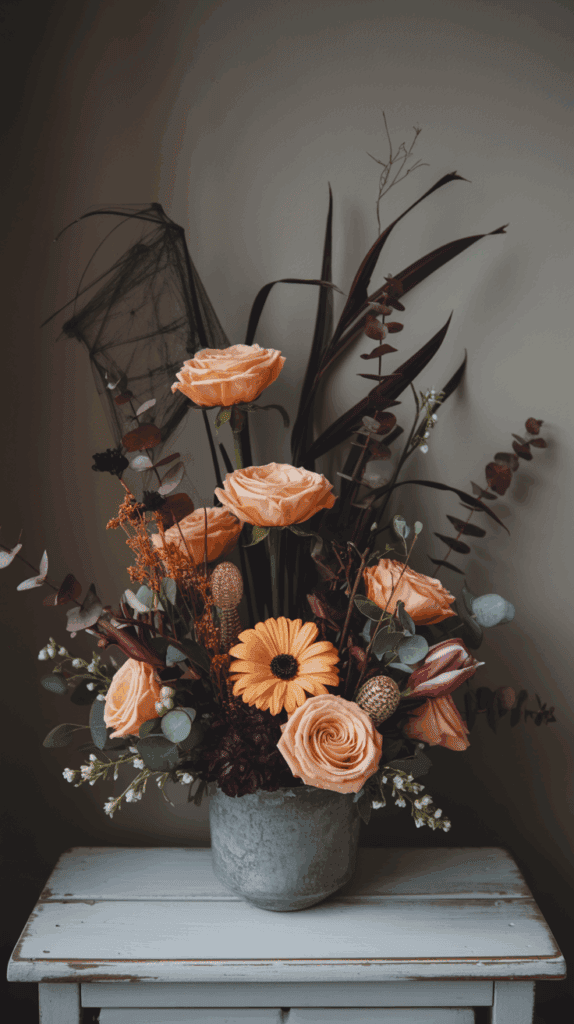 A floral arrangement featuring peach roses, orange gerbera daisies, and various dark purple and green foliage, set in a gray pot on a white distressed wooden table. A subtle black netting adds a touch of texture to the display.