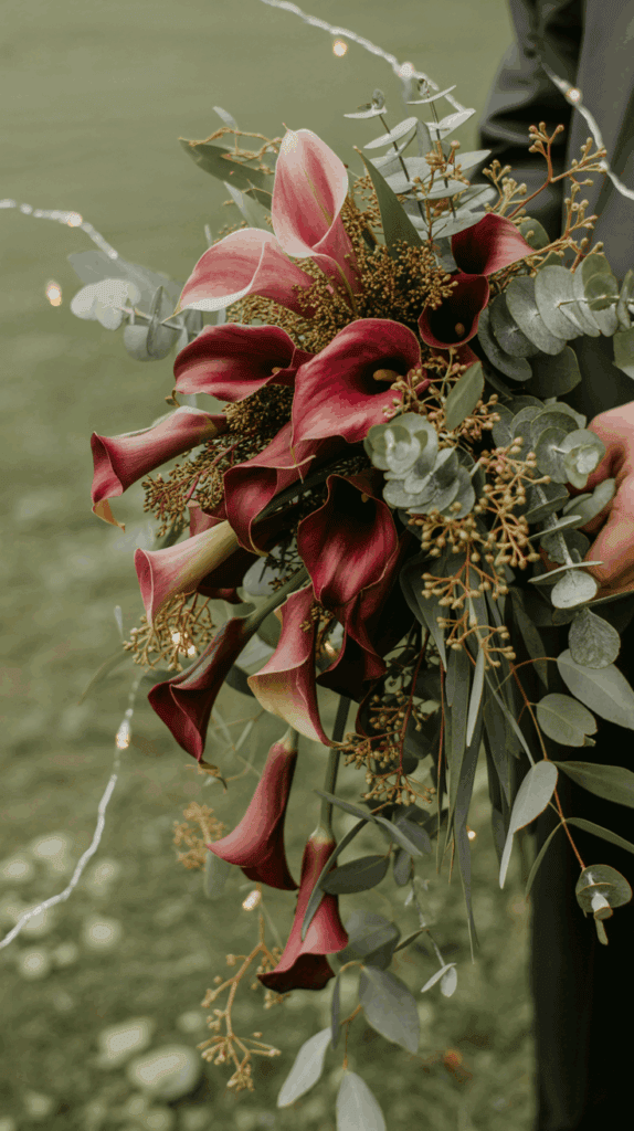 A bouquet featuring dark red calla lilies, eucalyptus leaves, and small green sprigs, held by a person, with tiny string lights woven through the arrangement.