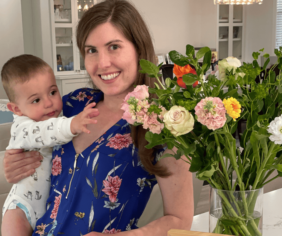 A woman holding a young child stands in a room with a vase of colorful flowers, including roses and carnations, on a table in front of them. The woman is smiling and wearing a blue floral dress, and the child is reaching towards the flowers.