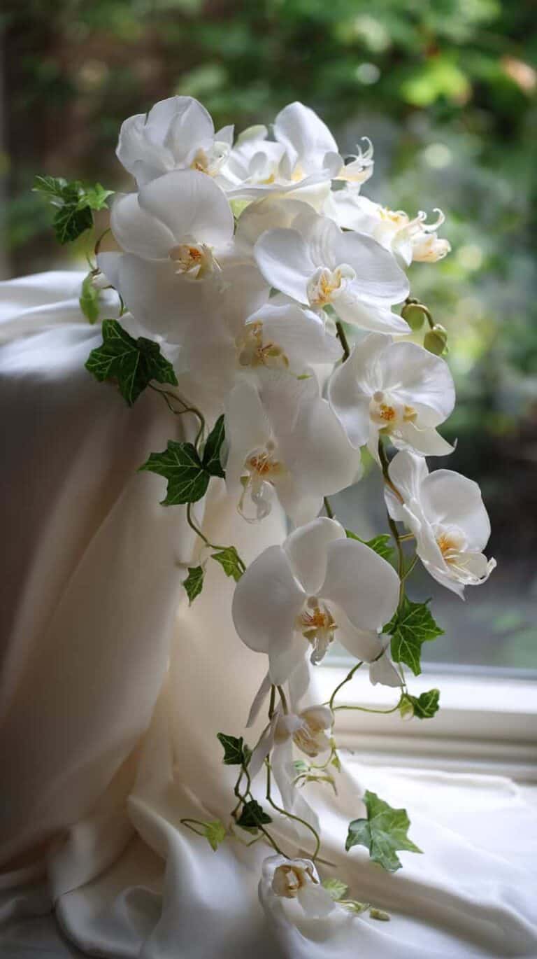 White orchids cascading down draped white fabric with green leaves, set against a blurred garden background.